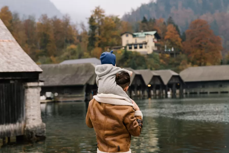 a man holding a baby on his shoulders by the water looking at fall trees